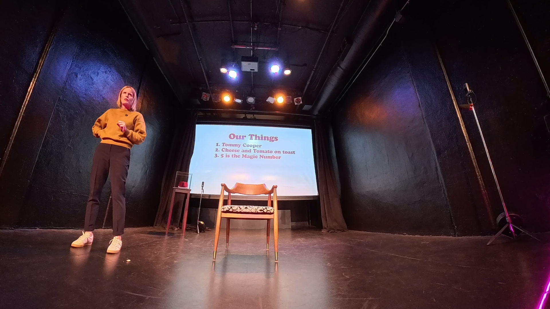 Performer on a black-box stage beside a 'Our Things' projection during Tommy Cooper's Fez